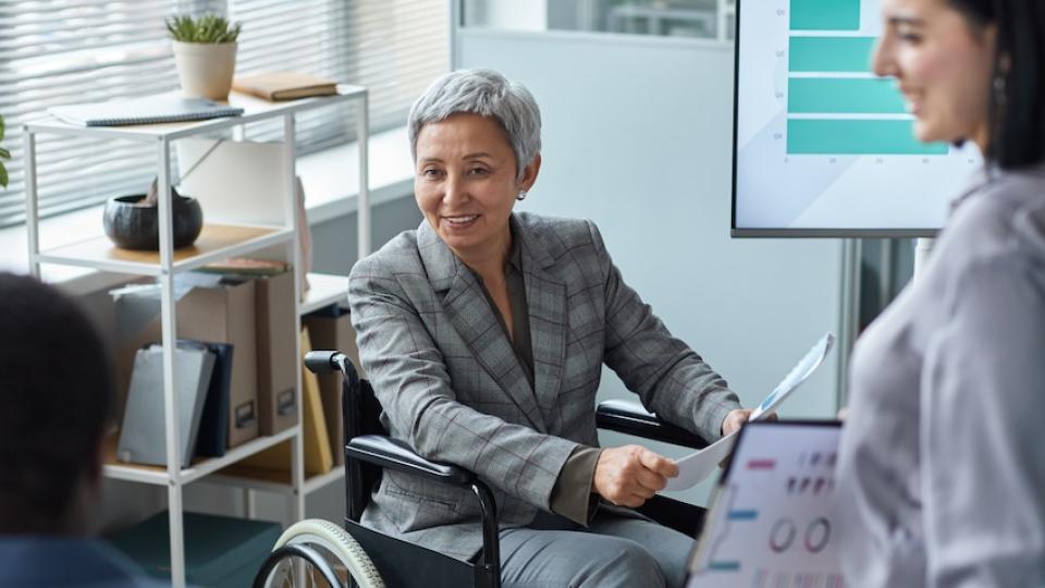 <p>An Adobe Stock image of a woman using a wheelchair and wearing a grey business suit meets with work colleagues.</p> A woman using a wheelchair and wearing a grey business suit meets with work colleagues.